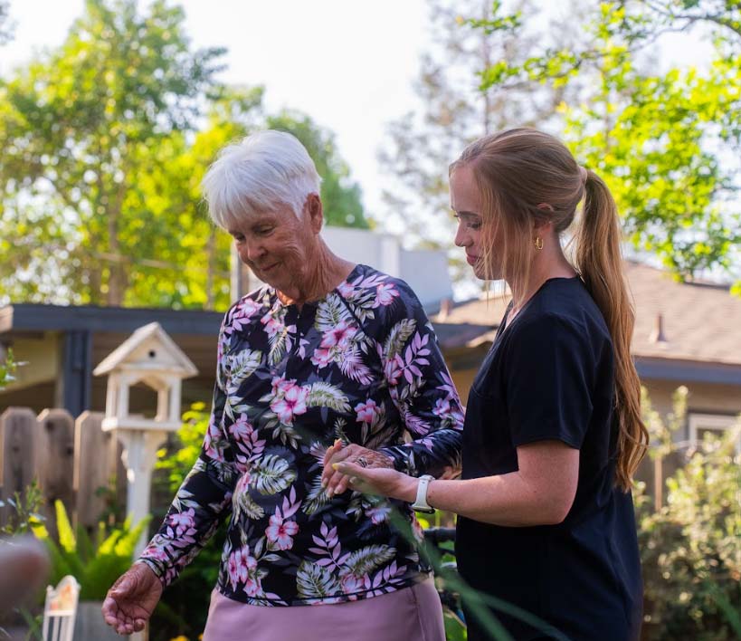A compassionate Amdal In Home Care in atascadero, ca caregiver providing gentle ambulation support to a senior woman in a sunny outdoor garden setting. The caregiver, dressed in professional black scrubs, holds the senior’s hand to ensure safety and stability during their walk. This image highlights our commitment to promoting independence and physical activity through personalized senior care.