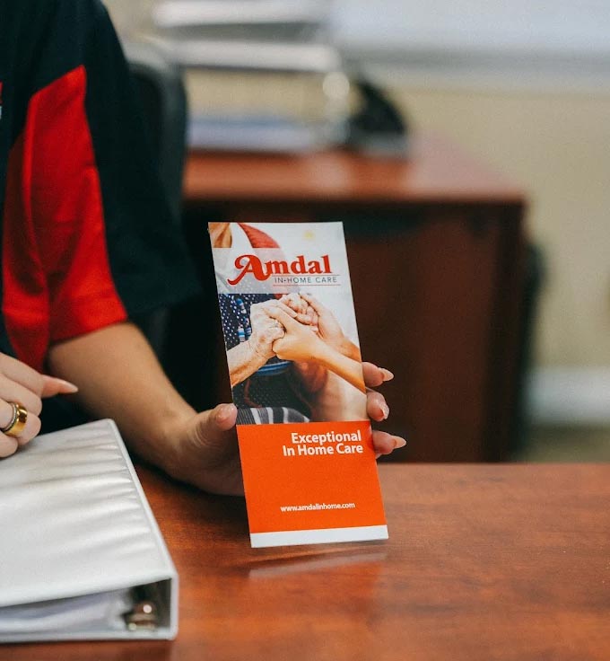 An Amdal In-Home Care team member holding an "Exceptional In Home Care" brochure on a wooden desk, highlighting professional senior care services