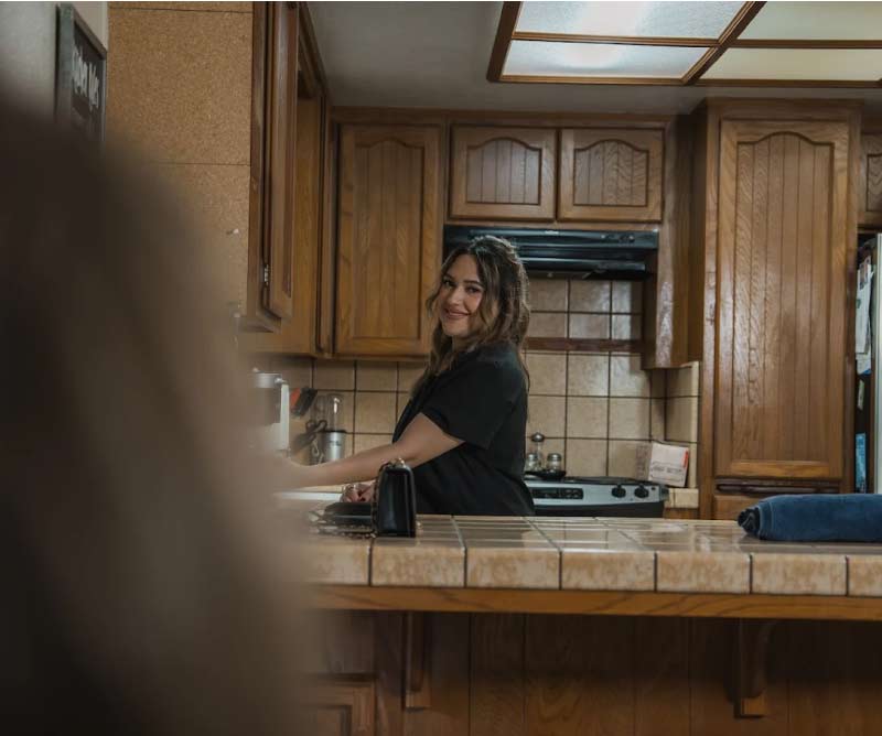 A smiling Amdal In-Home Care caregiver looking back while preparing a meal in a traditional home kitchen with wooden cabinetry