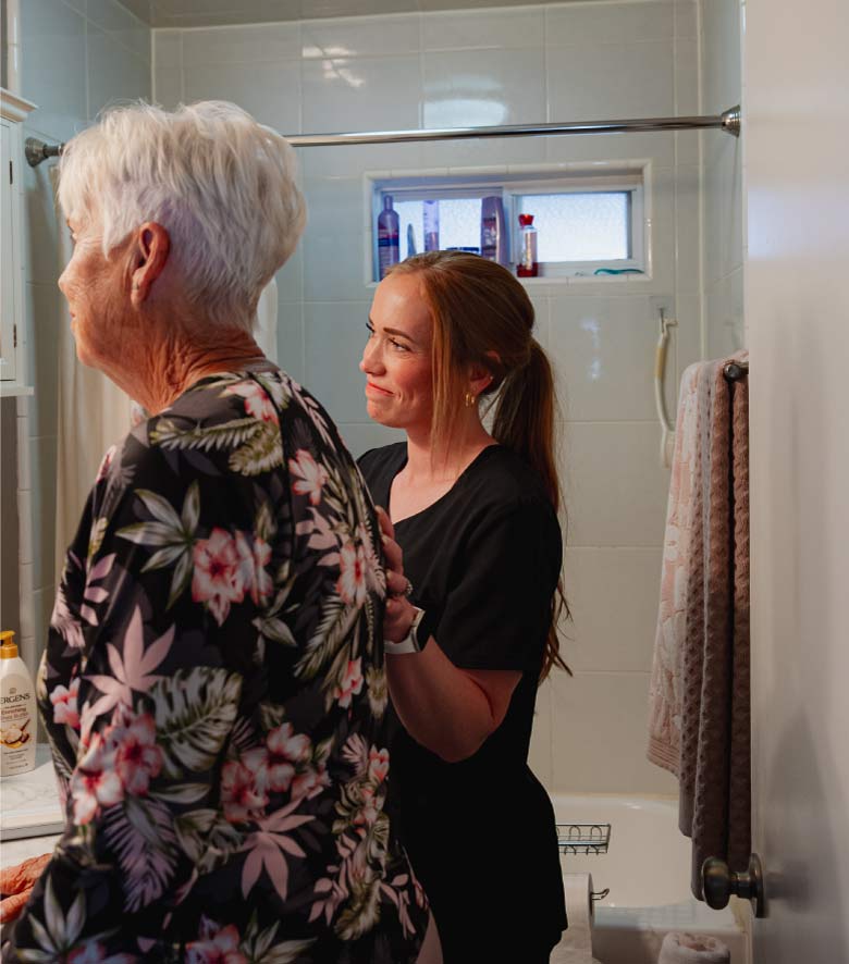 A compassionate caregiver providing personal care and grooming assistance to a senior woman in a brightly lit bathroom to ensure hygiene and safety