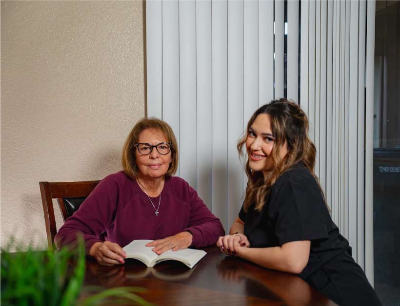 A friendly caregiver sitting at a table with a senior woman who is reading a book, demonstrating companionship and personalized in home care in porterville, ca