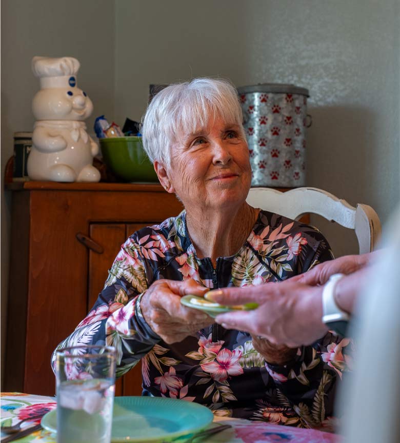 A senior woman in a floral shirt smiling warmly while an Amdal In-Home Care caregiver assists with meal preparation and serving at a dining table