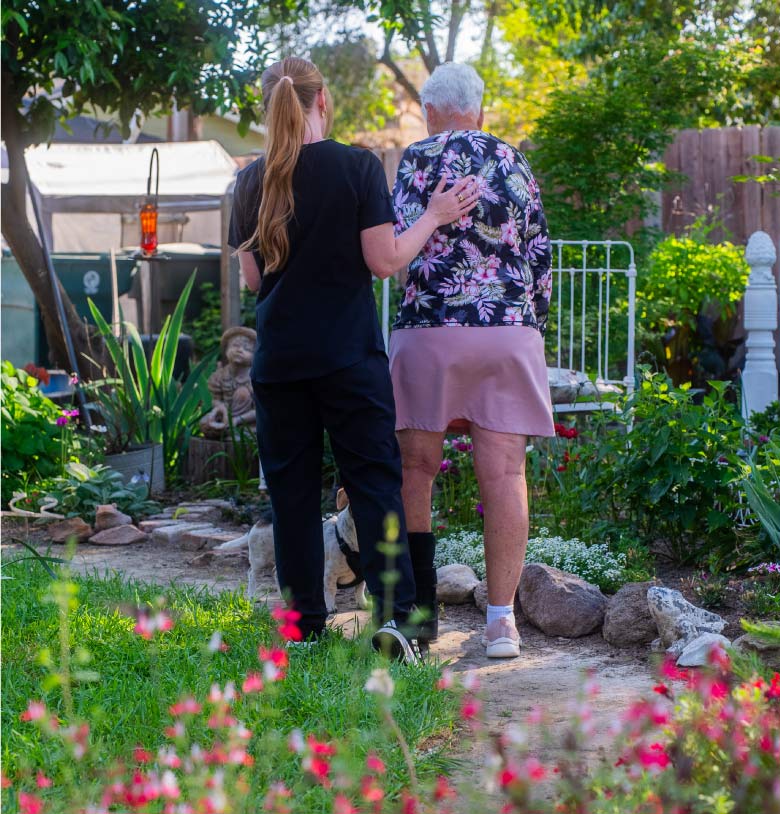 A caregiver providing mobility and ambulation support to a senior woman while walking through a garden to encourage physical activity and independence
