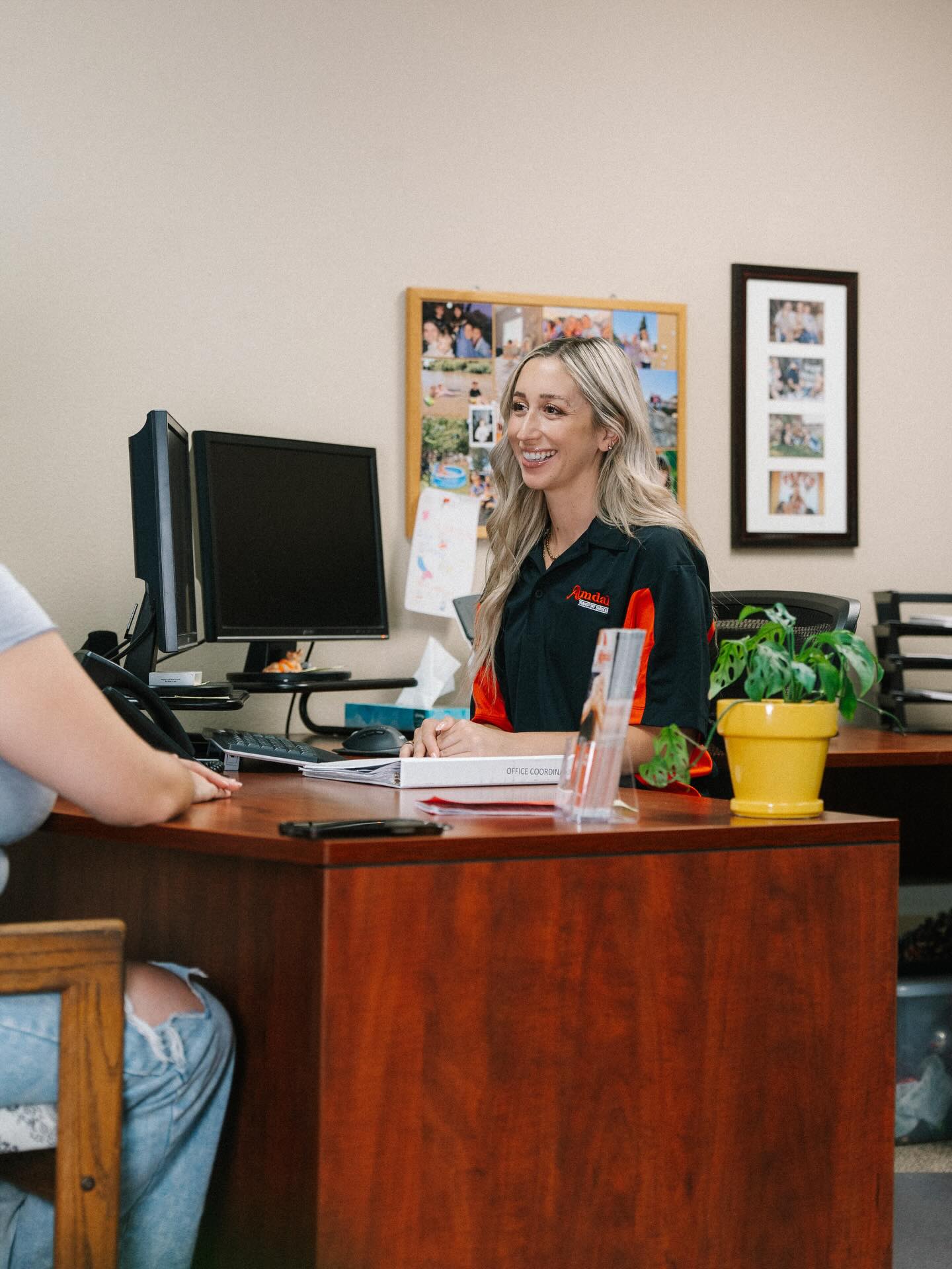 A friendly Amdal In-Home Care office coordinator with blonde hair and a black and orange polo shirt sits at a wooden desk. She is smiling warmly while assisting a client, exemplifying the compassionate and professional atmosphere of the Atascadero office. On the desk are dual monitors, a "Care Coordinator" binder, and a vibrant yellow potted plant, with framed photos on the wall behind her creating a welcoming, community-focused environment.